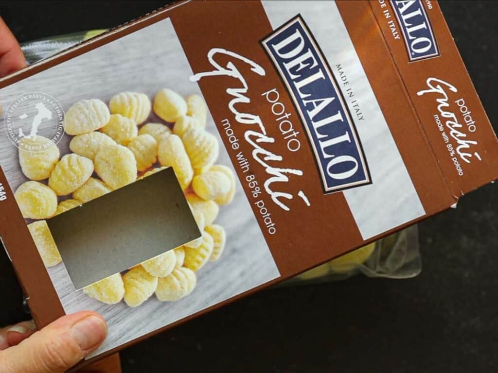 A person holds a brown DeLallo Potato Gnocchi box with a clear window showing gnocchi inside. The box text reads Made with 85% potato. The box is placed on a dark surface.